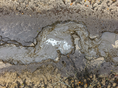 Aerial view of a small muddy stream flowing through wet textured soil with swirling water patterns and patches of dry earth in Latvia.