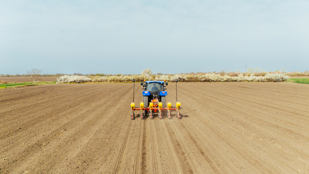 Aerial view of a blue tractor planting seeds in the brown earth with a backdrop of blooming trees under the muted sky Sremska Mitrovica Vojvodina Serbia. Aerial view of a blue tractor planting seeds in the brown earth with a backdrop of blooming trees under the muted sky Sremska Mitrovica Vojvodina Serbia.