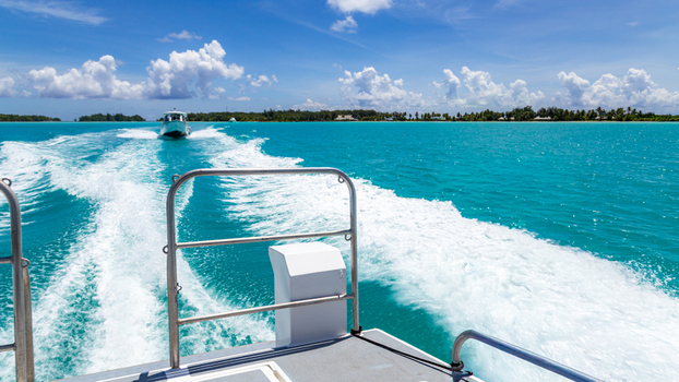 View of a boat wake trailing through vibrant turquoise lagoon waters with a distant boat and tropical coastline under a bright blue sky in Bora Bora Leeward Islands French Polynesia.