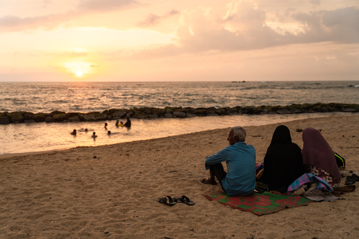 Sri Lanka 14 January 2026 View of people sitting on a sandy beach and others swimming in the ocean during a golden sunset with cloudy skies.