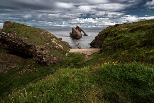 View of the Bow Fiddle Rock sea arch from the grassy cliffs under a dramatic cloudy sky in Scotland.