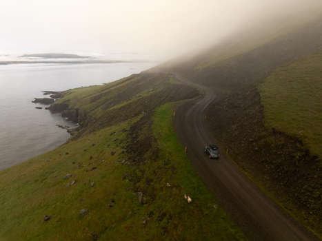 Aerial view of a car driving along a winding road on the green grassy slopes of the Eastern Region coastline in Iceland. Aerial view of a car driving along a winding road on the green grassy slopes of the Eastern Region coastline in Iceland.