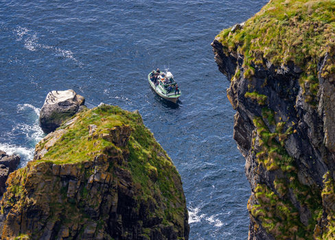 Aerial view of a small boat carrying passengers navigating through deep blue waters between steep grass covered rocky cliffs in Runde More og Romsdal Norway.