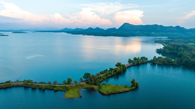 Aerial view of Upper Vaitarana Dam surrounded by lush green landscapes calm blue waters and distant hills under a soft sunset sky in Ondli Maharashtra India.