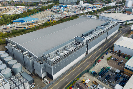 Aerial view of a large industrial data center facility featuring rooftop cooling units an adjacent parking lot and surrounding infrastructure in the United Kingdom. Aerial view of a large industrial data center facility featuring rooftop cooling units an adjacent parking lot and surrounding infrastructure in the United Kingdom.