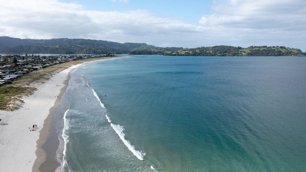 Aerial view of Omaha Beach with white sand turquoise water and coastal residential houses under a cloudy sky in Omaha Auckland New Zealand.