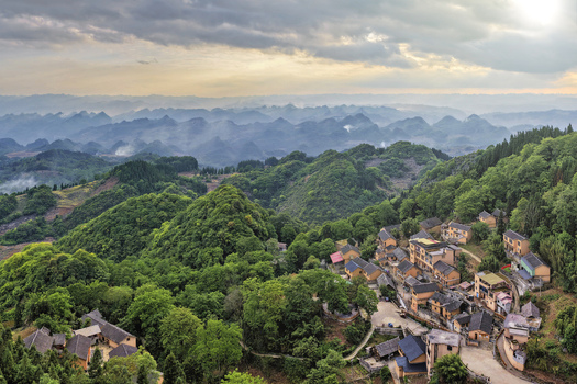 Aerial view of traditional houses in a remote mountain village surrounded by lush green karst peaks and misty valleys under a cloudy sky in Gaoxing Village Guizhou China.