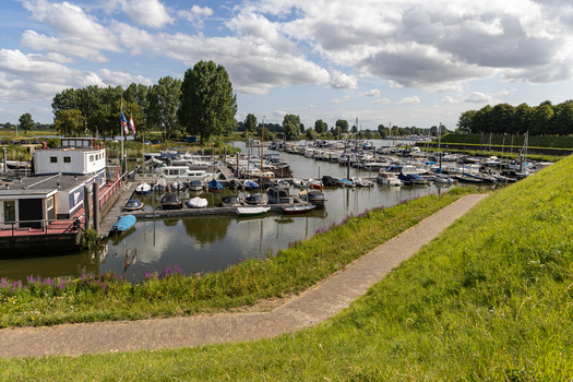 View of a marina with various boats docked along the pier surrounded by lush green grass and trees under a blue sky with clouds in Heusden North Brabant Netherlands.