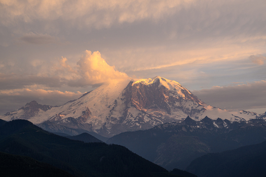 View of Mount Rainier at sunset with golden light on its snowy peak and surrounding clouds set against dark forested mountain ridges in Pierce County Washington United States. View of Mount Rainier at sunset with golden light on its snowy peak and surrounding clouds set against dark forested mountain ridges in Pierce County Washington United States.