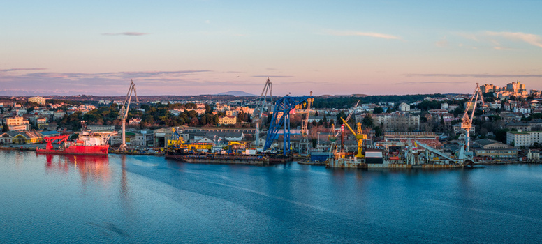 Aerial view of Uljanik Shipyard with industrial cranes ships docked at the pier and the cityscape of Pula in the background under a soft sunset sky in Pula Croatia. Aerial view of Uljanik Shipyard with industrial cranes ships docked at the pier and the cityscape of Pula in the background under a soft sunset sky in Pula Croatia.