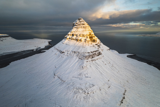 Aerial view of snow covered Kirkjufell mountain basks in the soft glow of the setting sun contrasting sharply with the dark moody sky above Kirkjufell Grundarfjar&eth;arb&aelig;r Iceland.