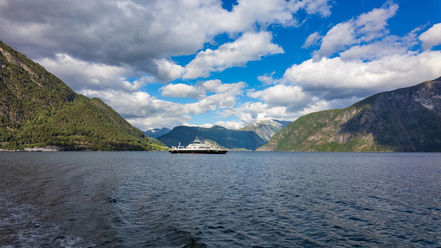 View of a ferry crossing the calm waters of Sunnylvsfjorden surrounded by steep green mountains under a cloudy sky More og Romsdal Norway.