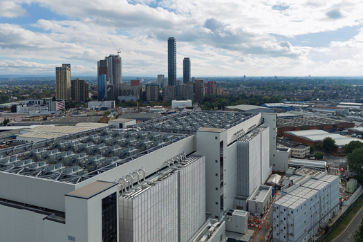 Aerial view of a large modern data center with extensive rooftop cooling infrastructure set against a distant urban skyline under a cloudy sky in the United Kingdom. Aerial view of a large modern data center with extensive rooftop cooling infrastructure set against a distant urban skyline under a cloudy sky in the United Kingdom.