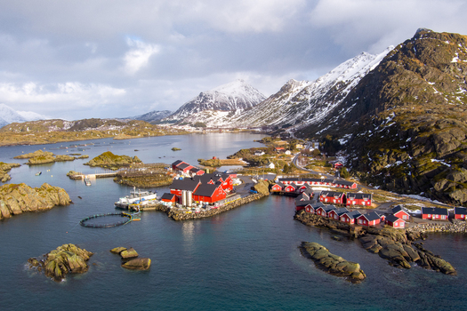 Aerial view of the Lofoten islands featuring red rorbuer cabins and a fish processing facility with circular pens surrounded by snow capped mountains and the sea in Nordland Norway.