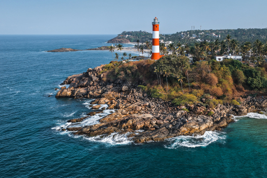 Aerial view of the red and white striped Vizhinjam Lighthouse standing on a rocky cliff overlooking the blue Arabian Sea and palm trees in Kovalam Kerala India. Aerial view of the red and white striped Vizhinjam Lighthouse standing on a rocky cliff overlooking the blue Arabian Sea and palm trees in Kovalam Kerala India.