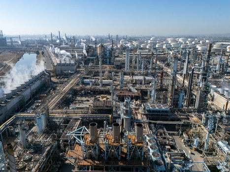 Aerial view of a large industrial oil refinery complex featuring extensive pipe networks storage tanks and cooling towers emitting steam in Carson California United States.