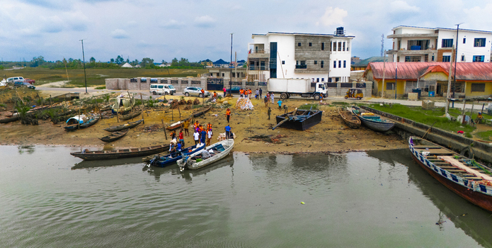 Aerial view of wooden boats docked along a muddy riverbank with people working near the water and modern buildings in the background in Rivers Nigeria.