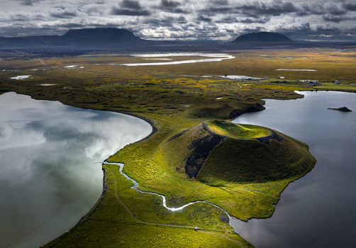 Aerial view of the Skutustadagigar pseudocraters at Lake Myvatn surrounded by green mossy landscape and calm water under a dramatic sky in Iceland.