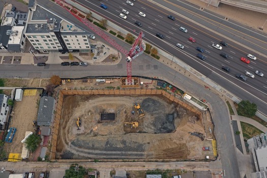 Aerial view of a construction site featuring a red tower crane and heavy machinery excavating the ground next to a busy highway in Denver Colorado United States. Aerial view of a construction site featuring a red tower crane and heavy machinery excavating the ground next to a busy highway in Denver Colorado United States.