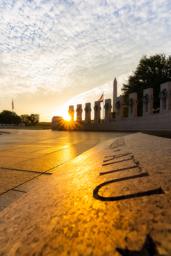 View of the World War II Memorial at sunrise with a sunburst effect over the stone pillars and the Washington Monument in the distance in Washington District of Columbia United States. View of the World War II Memorial at sunrise with a sunburst effect over the stone pillars and the Washington Monument in the distance in Washington District of Columbia United States.