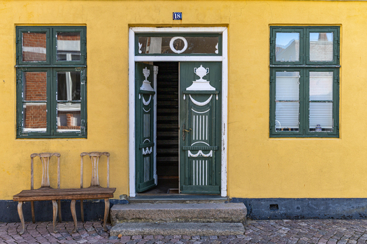 View of a traditional yellow house facade with an open green door and a wooden bench on a cobblestone street in Ribe Denmark. View of a traditional yellow house facade with an open green door and a wooden bench on a cobblestone street in Ribe Denmark.