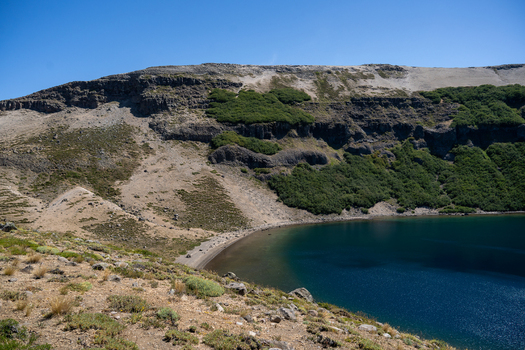 View of the crater lake at Volcan Batea Mahuida with rocky slopes and green vegetation under a clear blue sky in Alumine Neuquen Argentina. View of the crater lake at Volcan Batea Mahuida with rocky slopes and green vegetation under a clear blue sky in Alumine Neuquen Argentina.