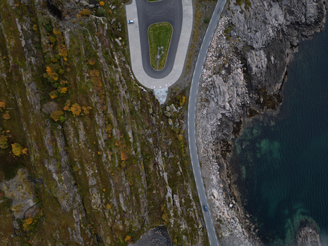 Aerial view of a coastal road winding between steep rocky cliffs with autumn foliage and the clear turquoise sea in Henningsvaer Nordland Norway.