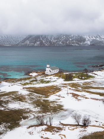 Aerial view of a solitary white church standing resiliently against the backdrop of snow dusted mountains and turquoise waters Gimsøysand Nordland Norway. Aerial view of a solitary white church standing resiliently against the backdrop of snow dusted mountains and turquoise waters Gimsøysand Nordland Norway.