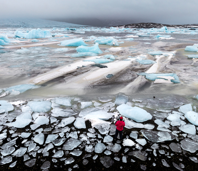 Aerial view of the stark ice strewn landscape meeting the turbulent ocean as a lone figure in red stands amidst the fragmented ice &Ouml;r&aelig;fi Iceland.