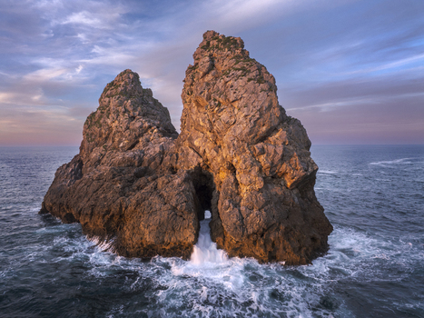 Aerial view of a rocky sea stack with waves crashing against the stone formation in the Cantabrian Sea under a soft sunset sky in Nueva Asturias Spain.