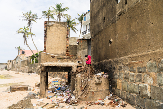 Sri Lanka 05 February 2026 View of a woman in a pink dress standing by a weathered stone wall and debris on a sandy beach with palm trees.