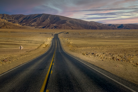 View of a long asphalt road stretching through the arid desert landscape of Death Valley National Park s backcountry under a soft sunset sky in Death Valley California United States.