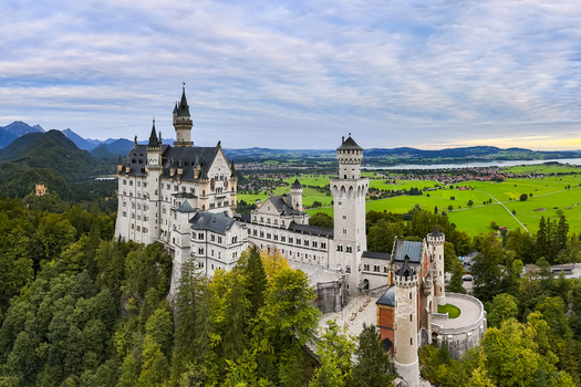 Aerial view of Neuschwanstein Castle standing majestically amidst verdant forests against a backdrop of distant mountains and serene lakes Neuschwanstein Castle Bayern Germany. Aerial view of Neuschwanstein Castle standing majestically amidst verdant forests against a backdrop of distant mountains and serene lakes Neuschwanstein Castle Bayern Germany.