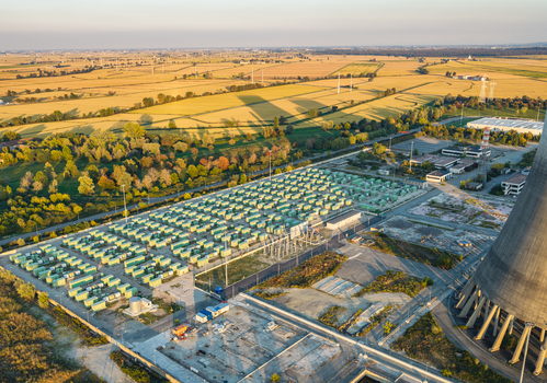 Aerial view of a large battery storage facility with rows of green containers situated beside industrial cooling towers and expansive golden farmland in Italy. Aerial view of a large battery storage facility with rows of green containers situated beside industrial cooling towers and expansive golden farmland in Italy.