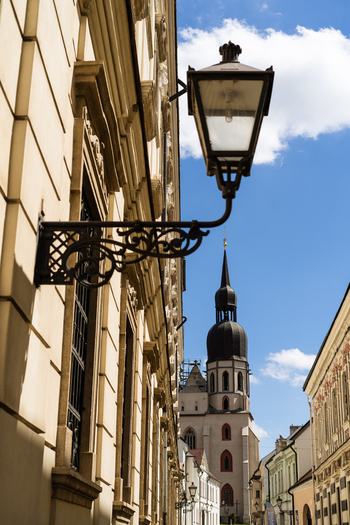 View of an ornate vintage street lamp mounted on a historic building facade leading towards the St. Nicholas Basilica tower under a blue sky in Trnava Trnava Region Slovakia. View of an ornate vintage street lamp mounted on a historic building facade leading towards the St. Nicholas Basilica tower under a blue sky in Trnava Trnava Region Slovakia.