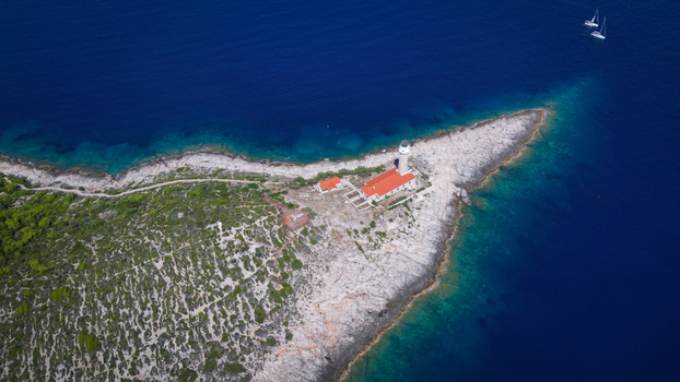 Aerial view of Stoncica lighthouse with its terracotta roof standing proud against the rugged coastline contrasted by the deep blue Adriatic Sea Vis Croatia.