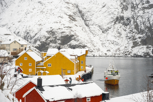 View of a fishing boat arriving at a dock in a snowy village with traditional yellow and red wooden houses nestled against dramatic mountains in Leknes Lofoten Norway.