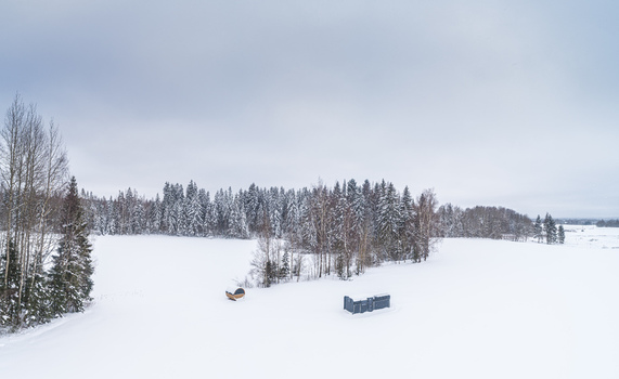 Aerial view of a snow covered field with a small wooden structure and a dark rectangular building surrounded by a dense forest of snow laden pine trees in Latvia. Aerial view of a snow covered field with a small wooden structure and a dark rectangular building surrounded by a dense forest of snow laden pine trees in Latvia.