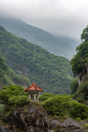 View of a traditional pavilion perched on a rocky outcrop amidst lush green mountains and misty cliffs in Taroko National Park Xiulin Township Hualien County Taiwan.