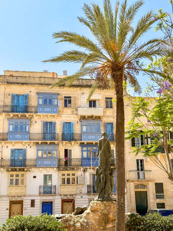 View of a statue stands proudly before a weathered building with blue balconies framed by a palm tree under a clear sky Valletta Malta Malta.