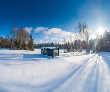 Aerial view of a modern dark colored cabin situated in a vast snow covered field surrounded by a forest of bare trees under a bright blue sky in Latvia. Aerial view of a modern dark colored cabin situated in a vast snow covered field surrounded by a forest of bare trees under a bright blue sky in Latvia.