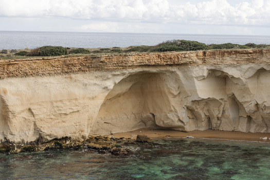 View of the rugged limestone cliffs and a natural sea cave overlooking the clear turquoise waters of the Mediterranean coast in Syracuse Sicily Italy.