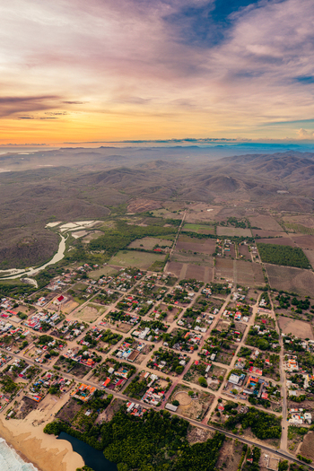 Aerial view of Punta Perula coastal town with grid streets lush vegetation and surrounding hills under a vibrant sunset sky in Perula Jalisco Mexico.