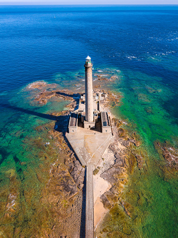 Aerial view of Gatteville Lighthouse standing on a rocky outcrop surrounded by clear turquoise and deep blue sea waters in Gatteville le Phare Normandie France.