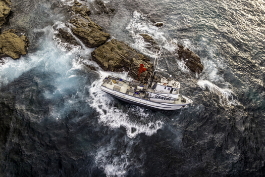 Aerial view of a fishing boat grounded on rocky outcrops near the headland with waves crashing against the hull in Bermagui NSW Australia.