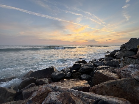 View of the sunset over the Pacific Ocean with waves crashing against a rocky shoreline under a sky with orange clouds and contrails in Oceanside California United States. View of the sunset over the Pacific Ocean with waves crashing against a rocky shoreline under a sky with orange clouds and contrails in Oceanside California United States.