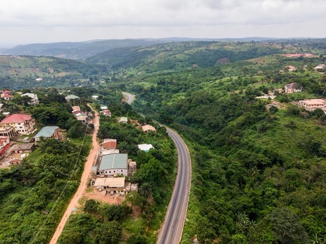 Aerial view of a winding asphalt road cutting through a lush green hilly landscape with scattered residential houses and dense vegetation in Ghana. Aerial view of a winding asphalt road cutting through a lush green hilly landscape with scattered residential houses and dense vegetation in Ghana.