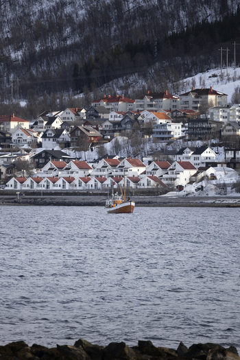 View of a traditional wooden fishing boat sailing on the water with snow covered hills and residential houses in the background in Tromso Troms Norway.