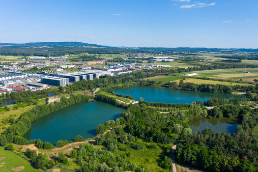 Aerial view of Sportinsel Forchheim Spazierweg und Spielplatz featuring blue lakes surrounded by lush green trees and industrial buildings in Forchheim Bayern Germany.