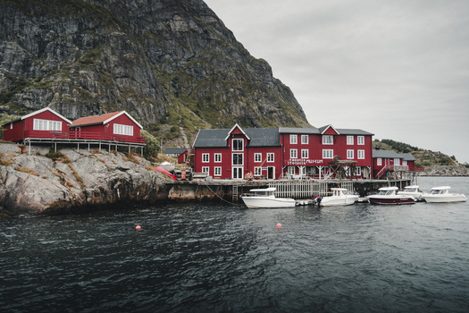 A Nordland Norway 04 September 2025 View of traditional red rorbu cabins and the fishing museum nestled against rocky cliffs by the sea with small motorboats moored at the pier.